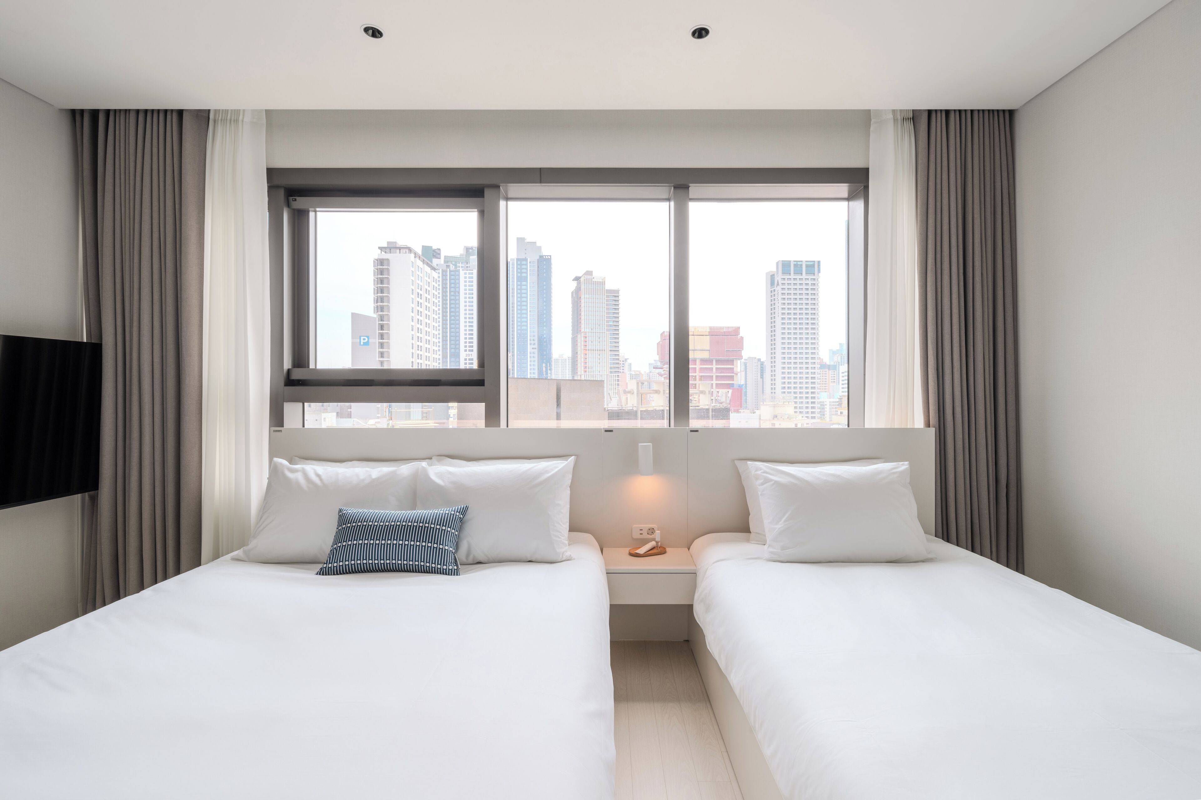 A hotel room with two beds, a large window with a city view, a flat-screen TV mounted on the wall, and a bedside table with a lamp.