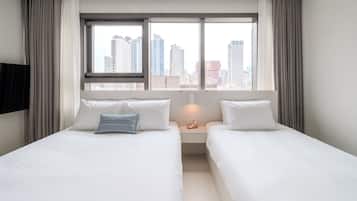 A hotel room with two beds, a large window with a city view, a flat-screen TV mounted on the wall, and a bedside table with a lamp.