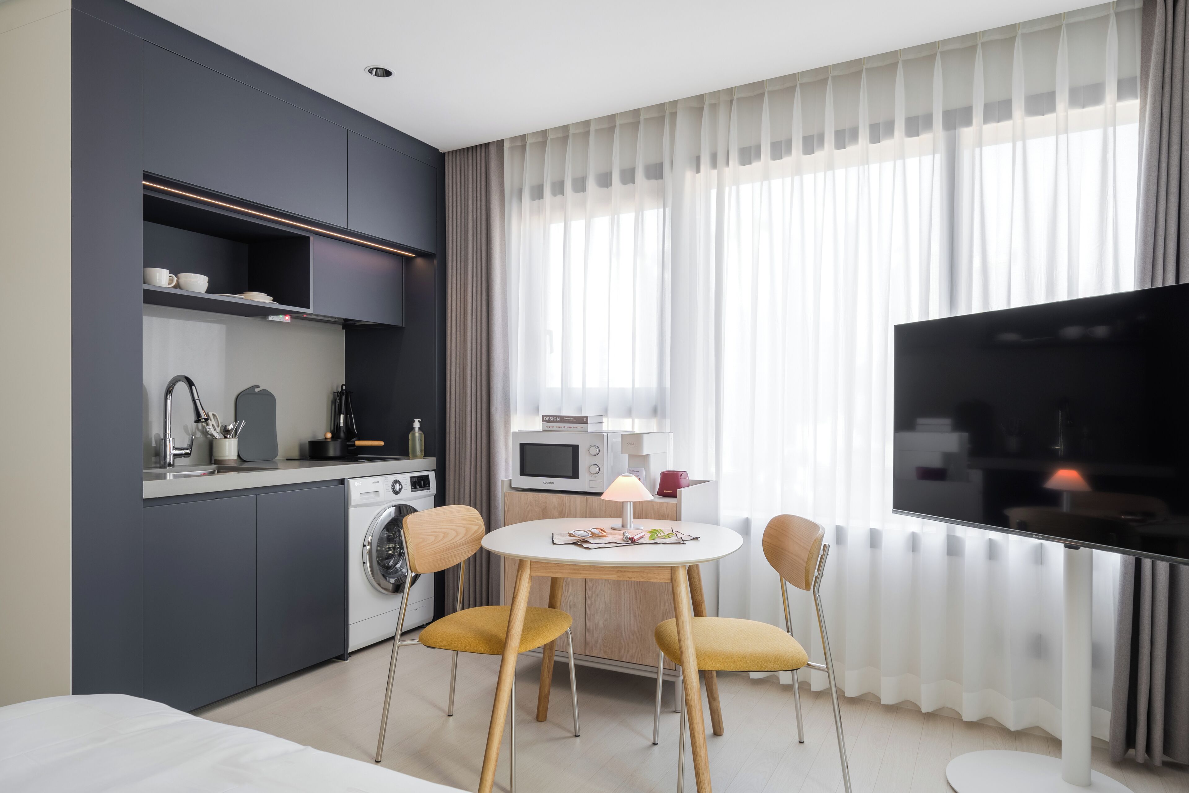 A modern hotel room with a dining area, kitchenette, and a large TV screen.