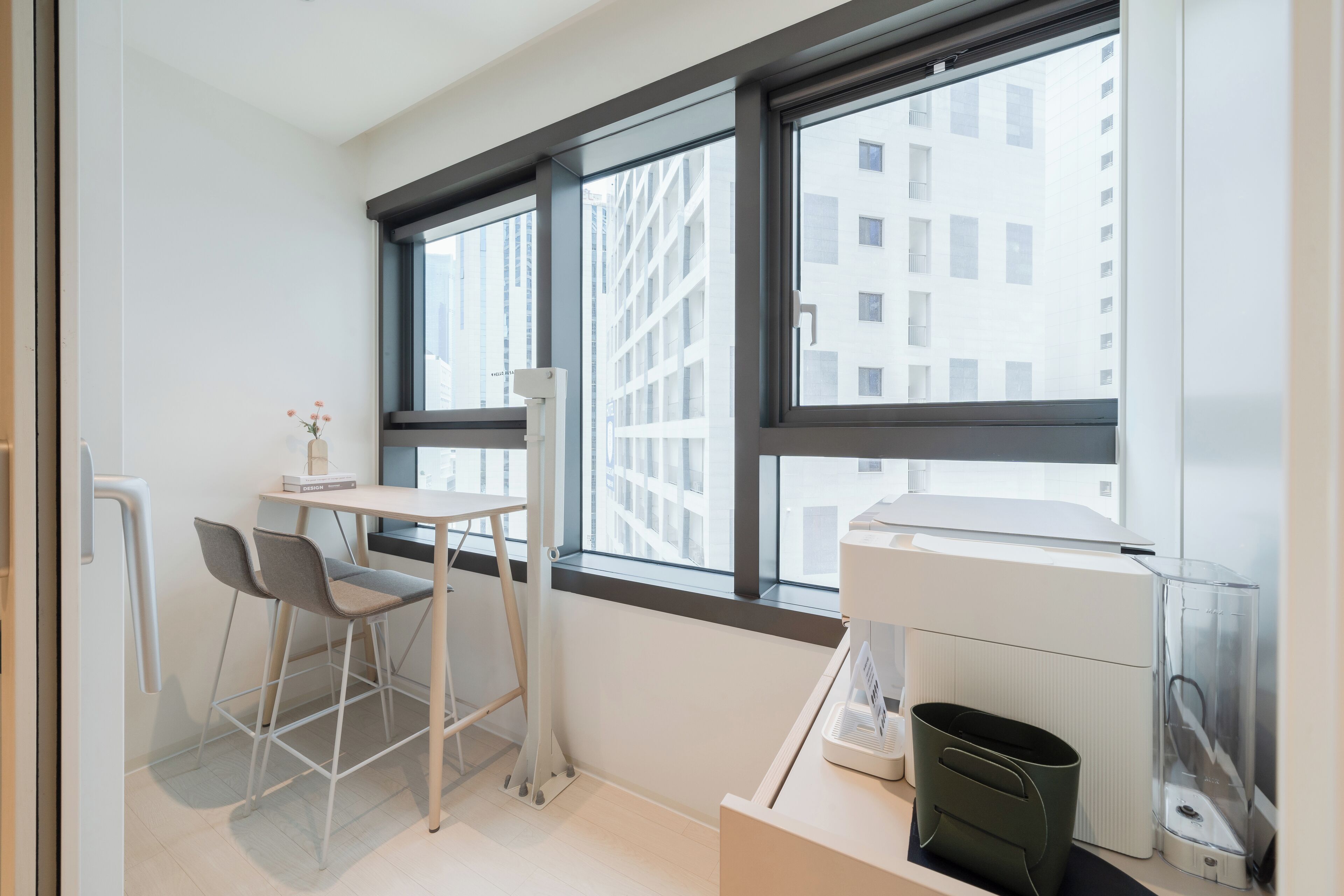 Une pièce moderne et bien éclairée, équipée d’un bureau, de deux chaises, d’un radiateur et de grandes fenêtres donnant sur un immeuble.