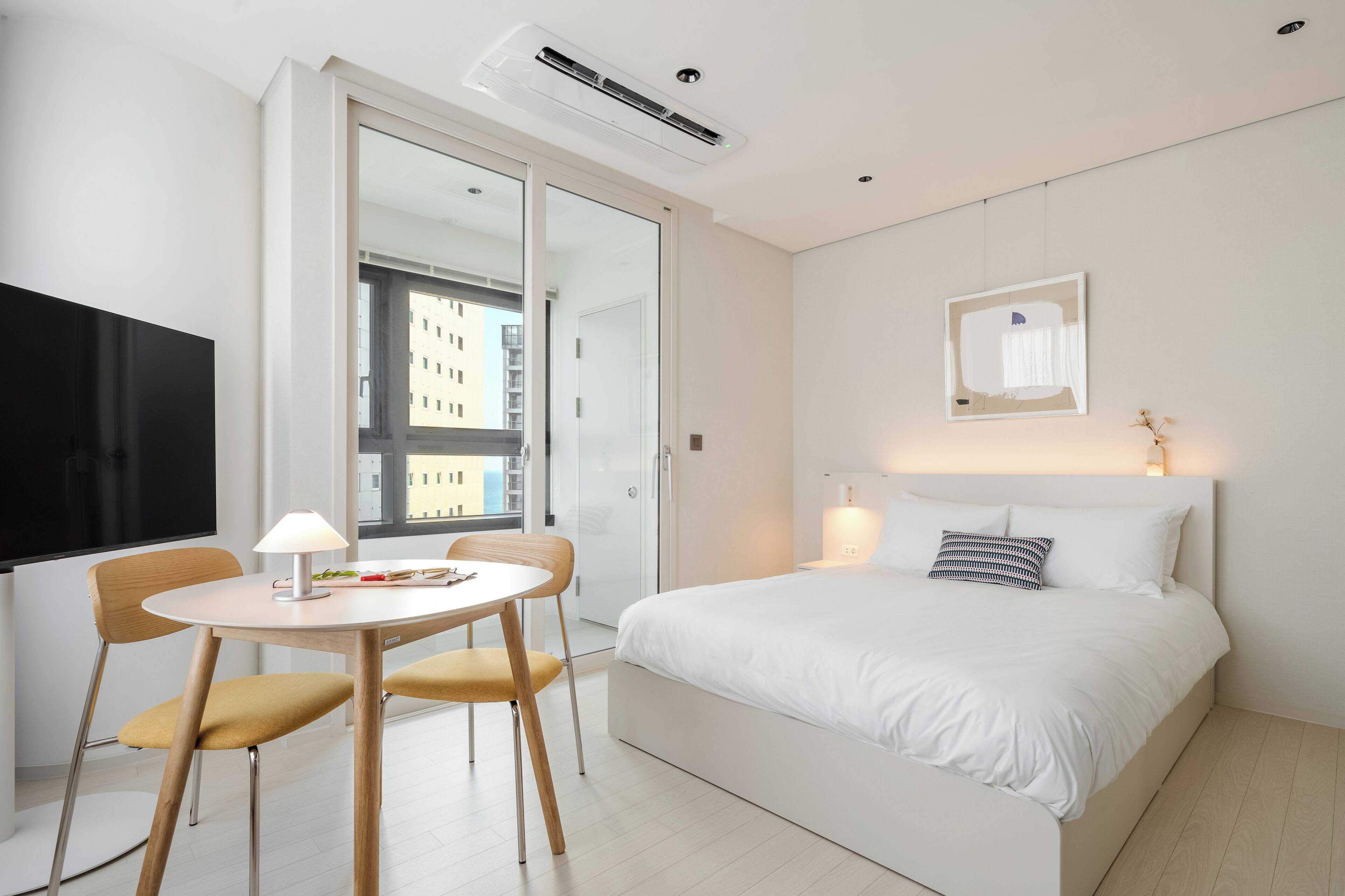 A modern hotel room with a bed, bedside table, lamp, and a view of the city through a large window.