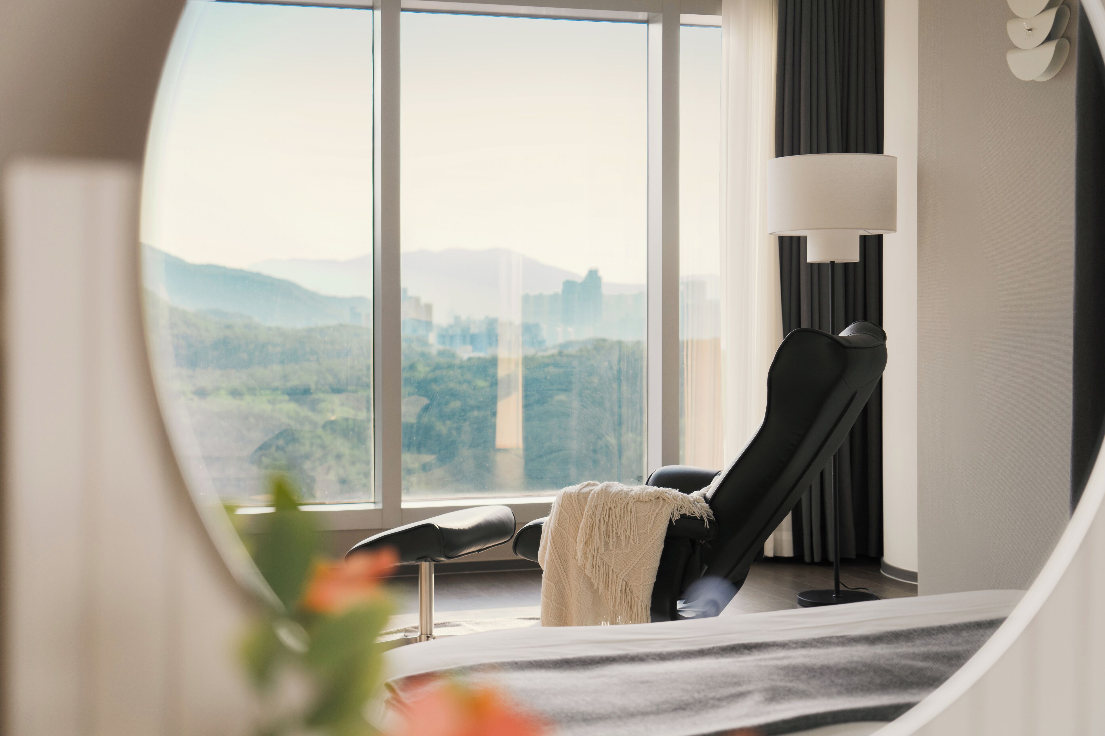 Una habitación moderna con un ventanal grande, un sillón reclinable negro, una manta blanca y una lámpara de pie.