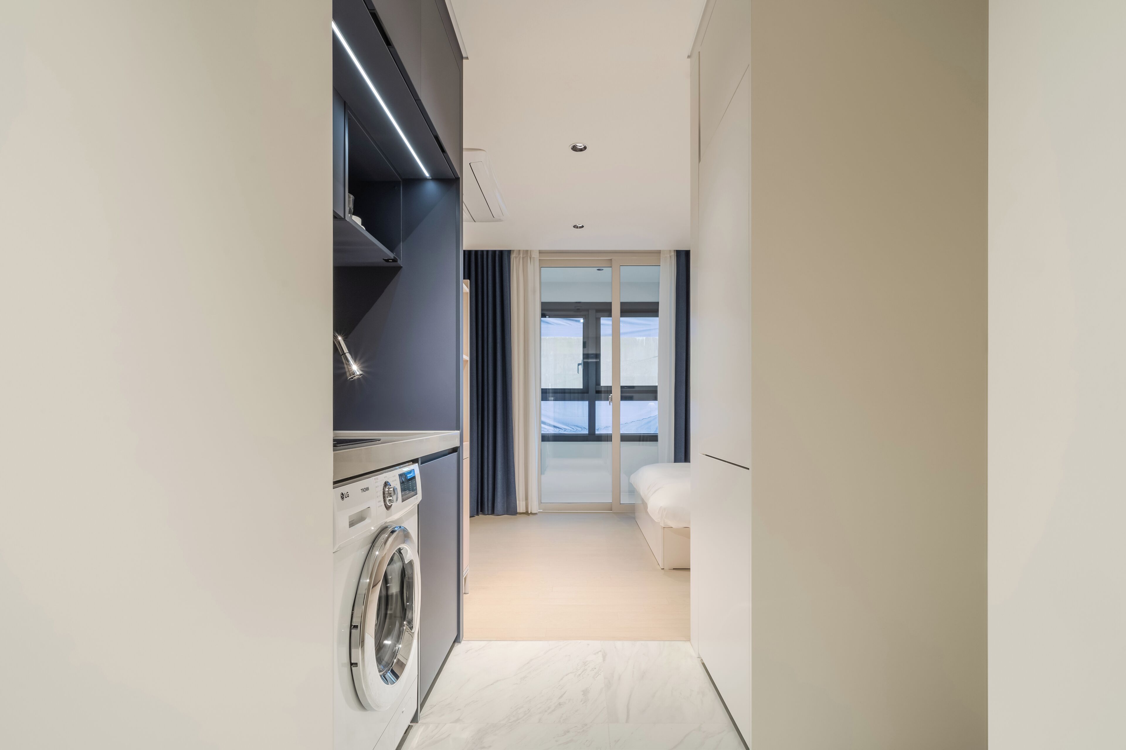 A modern hotel room with a washing machine, a bed, and a large window with curtains.