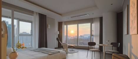 Habitación de hotel moderna con una cama grande, un escritorio, una silla, una mesita y vistas a la ciudad al atardecer.