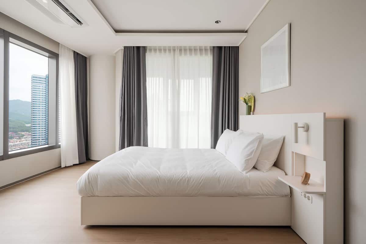 Ein modernes Schlafzimmer mit einem großen Bett, einem Nachttisch, einem Fenster mit Vorhängen und Blick auf die Stadt.
