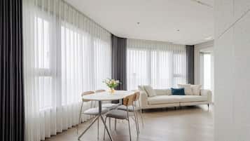 A modern living room with a dining area, featuring a sofa, a dining table with chairs, and large windows with sheer curtains.