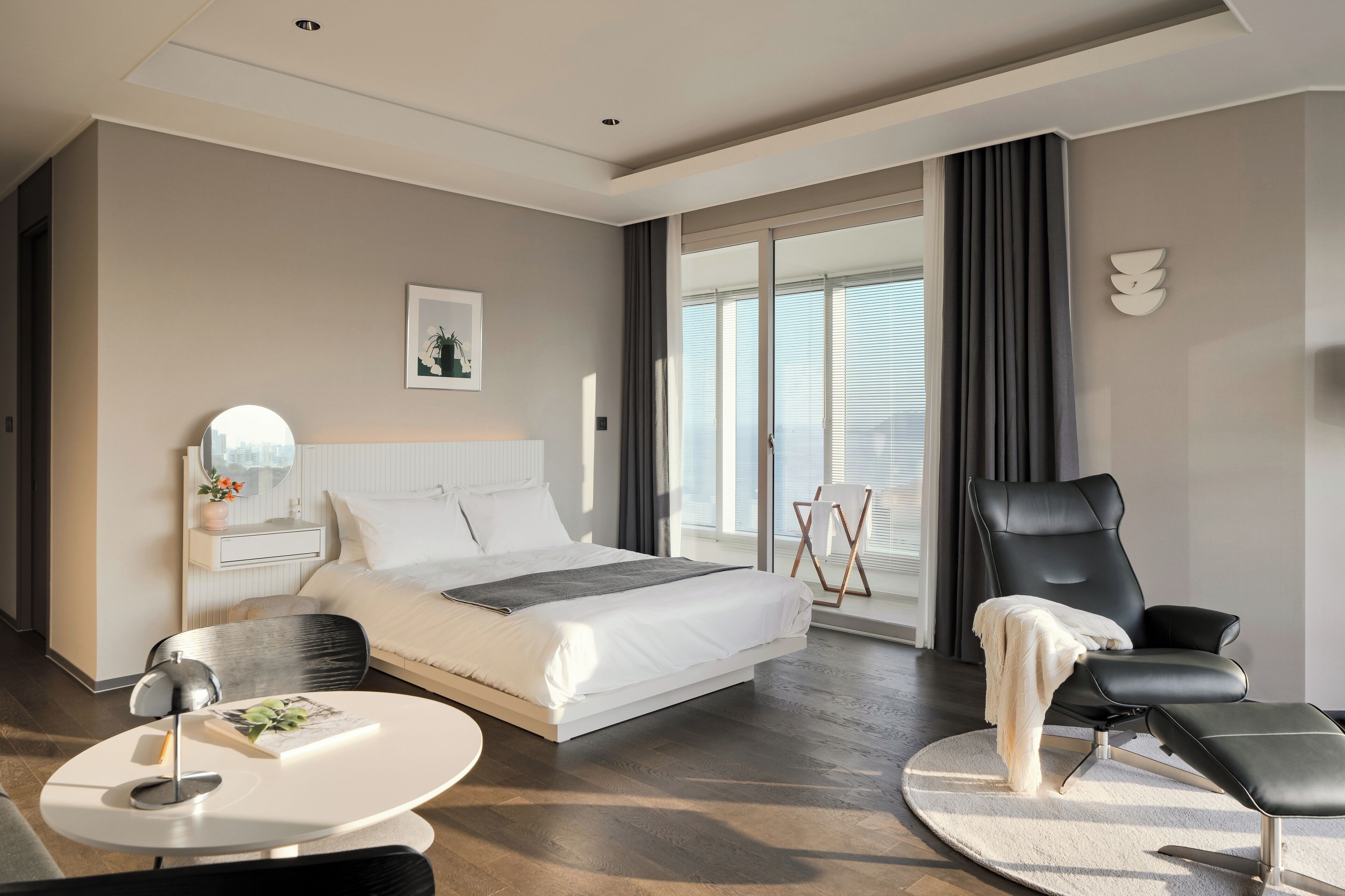 A modern hotel room with a large bed, a sitting area with a chair and ottoman, a round coffee table, and a view of the outdoors through a large window.