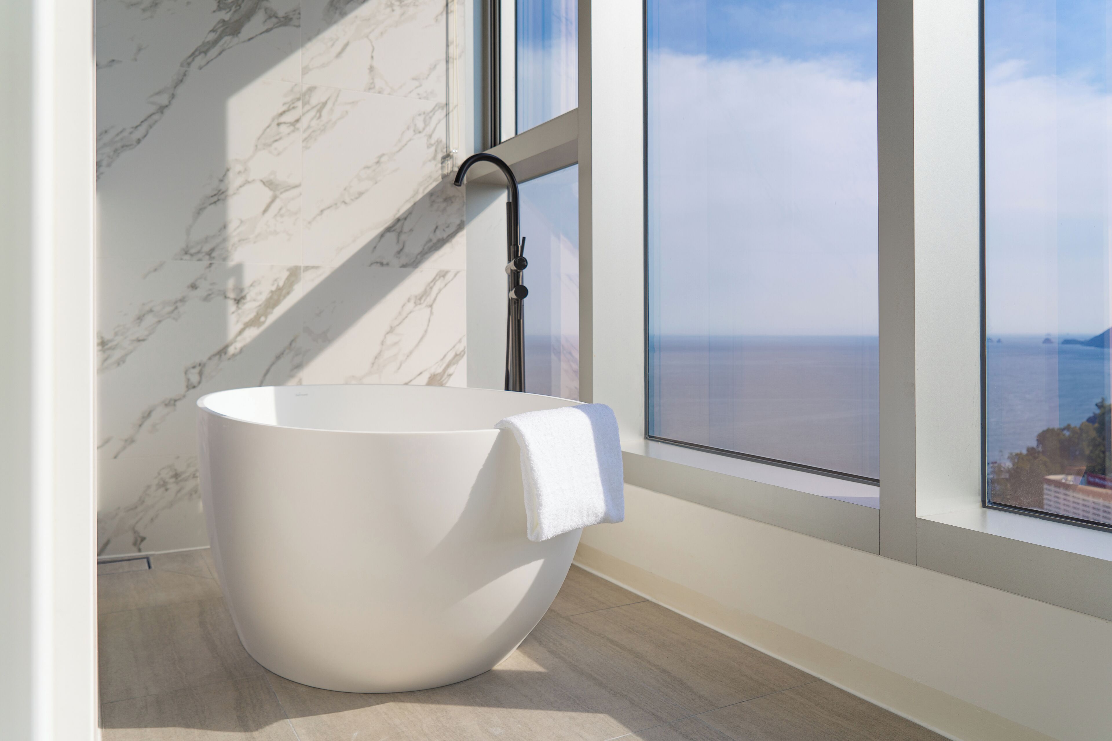 A modern bathroom with a freestanding bathtub, marble walls, and a large window offering a view of the sea.
