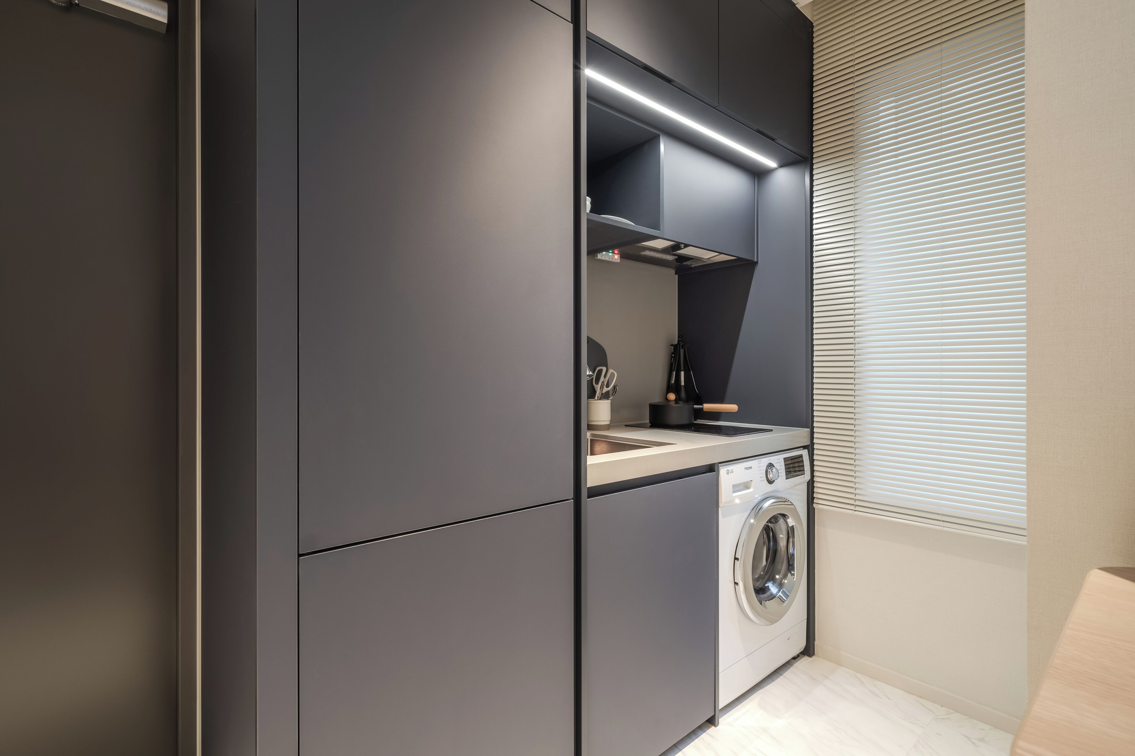 A compact kitchen with dark cabinetry, a built-in washing machine, and a window with blinds.
