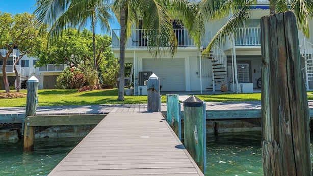 Un muelle de madera que lleva a una casa con garaje, rodeada de palmeras y un cielo azul despejado.