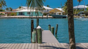 Une jetée en bois menant à une marina où sont amarrés des bateaux, un ciel bleu dégagé et des maisons en arrière-plan.