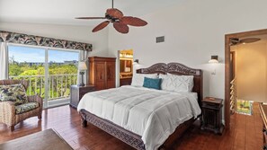 A bedroom with a large bed, a ceiling fan, a balcony with a view, and a wooden floor.