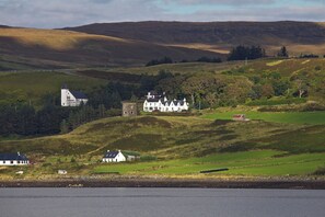 A coastal landscape with a prominent white building with multiple chimneys, surrounded by rolling hills and a body of water in the foreground. - Ascribs View (Uig)