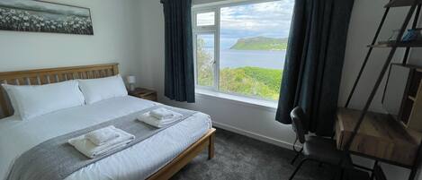 A bedroom with a large bed, a view of the sea, a window with curtains, a wooden headboard, a bedside table with a lamp, and a chair.