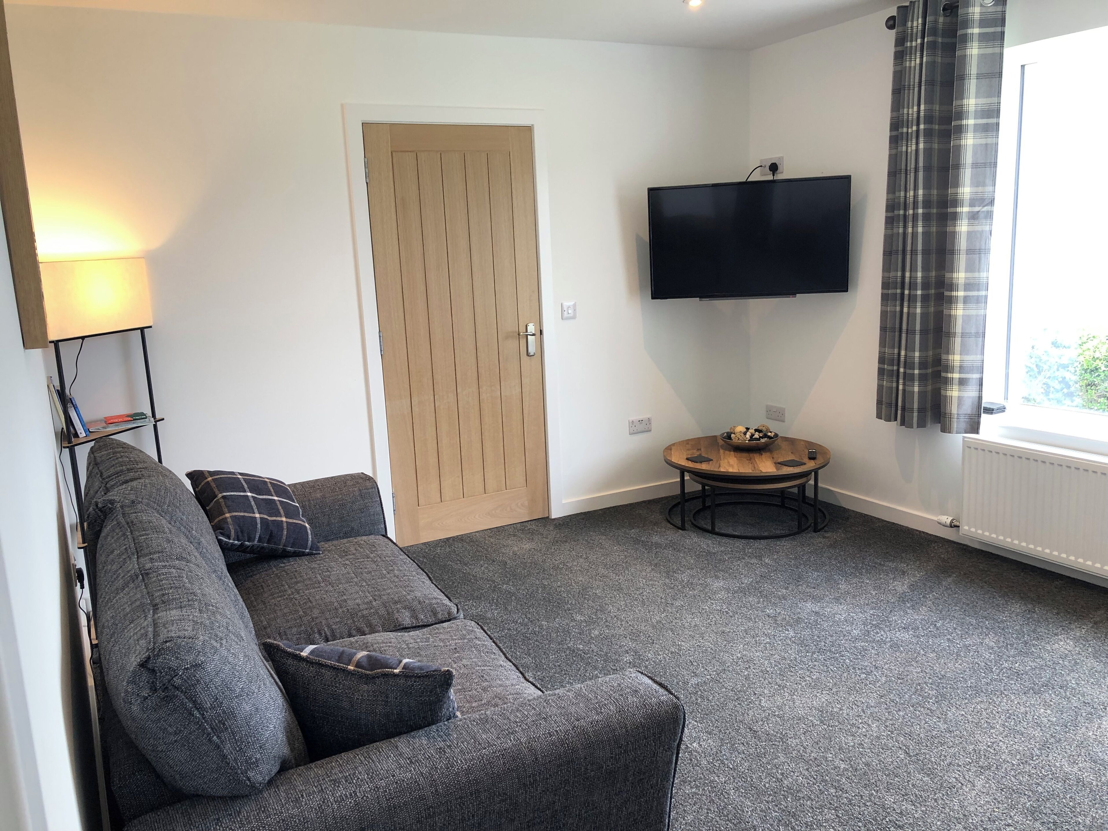 A living room with a grey sofa, a wooden door, a flat-screen TV, a small round table, and a window with curtains.