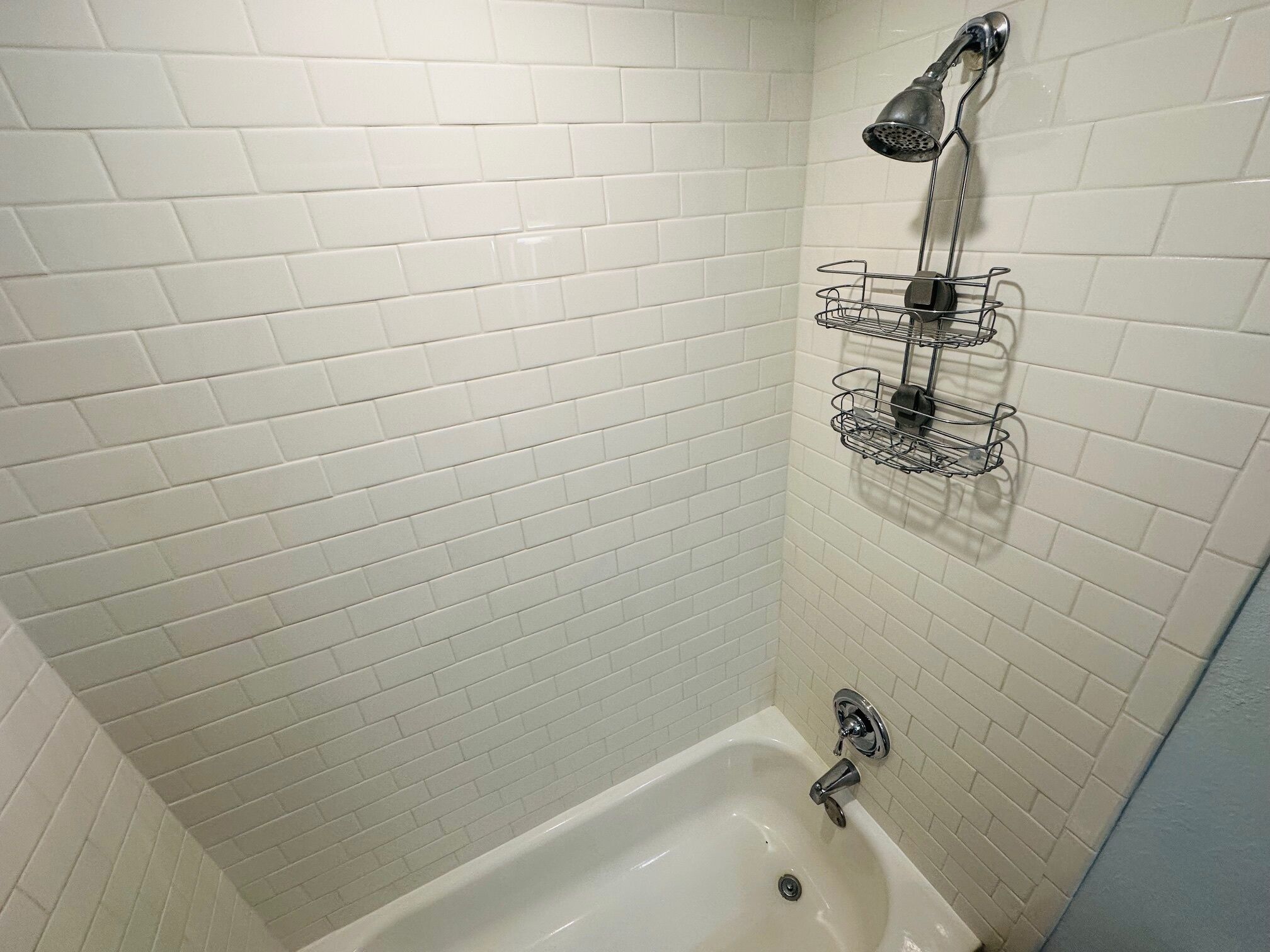 A bathroom with white tiled walls, a white bathtub, a showerhead, and a metal shelf with toiletries.