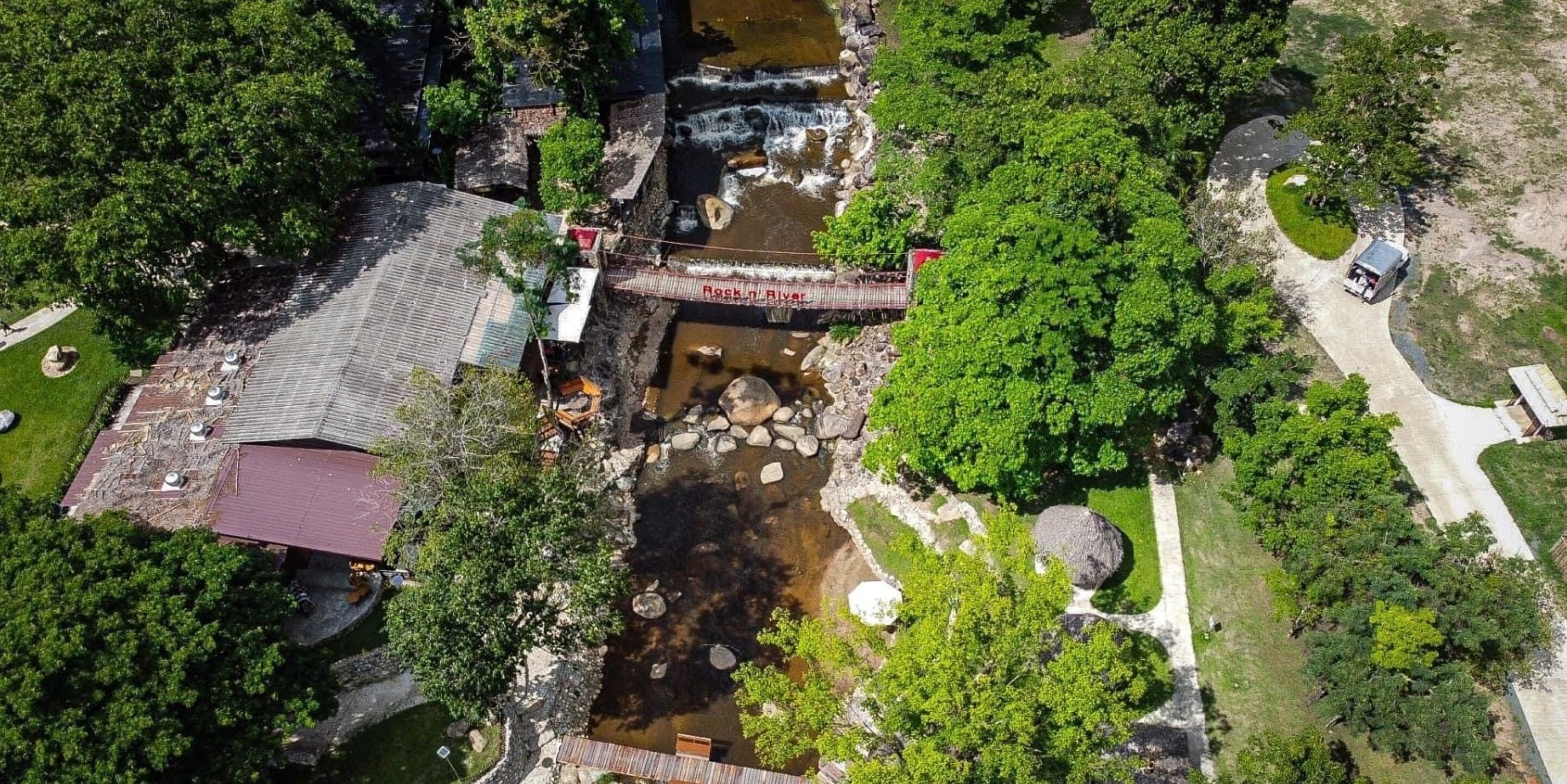 An aerial view of a riverside property with a wooden bridge, a small waterfall, and lush greenery.