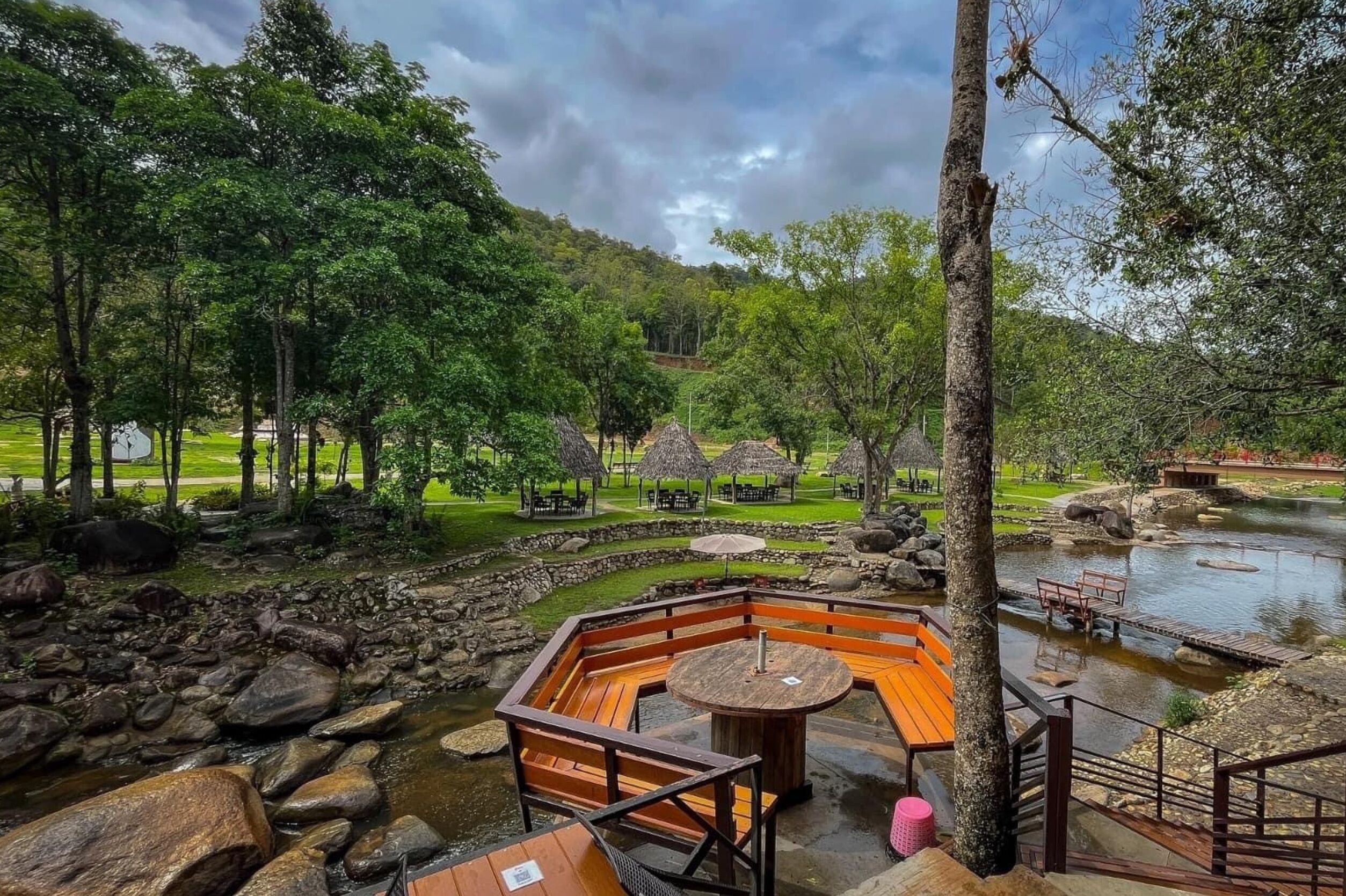 Een terras aan de rivier met houten zitplaatsen, een klein vijvertje en gebouwen met rieten daken.