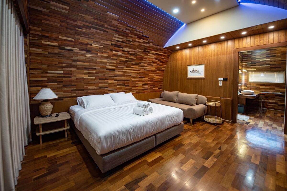 A modern bedroom with a wooden wall panel design, a large bed, a sofa, a small table, and a bathroom area.