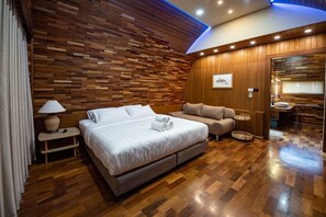 A modern bedroom with a wooden wall panel design, a large bed, a sofa, a small table, and a bathroom area. - Pang fan Rock & River Resort (Doi Saket)