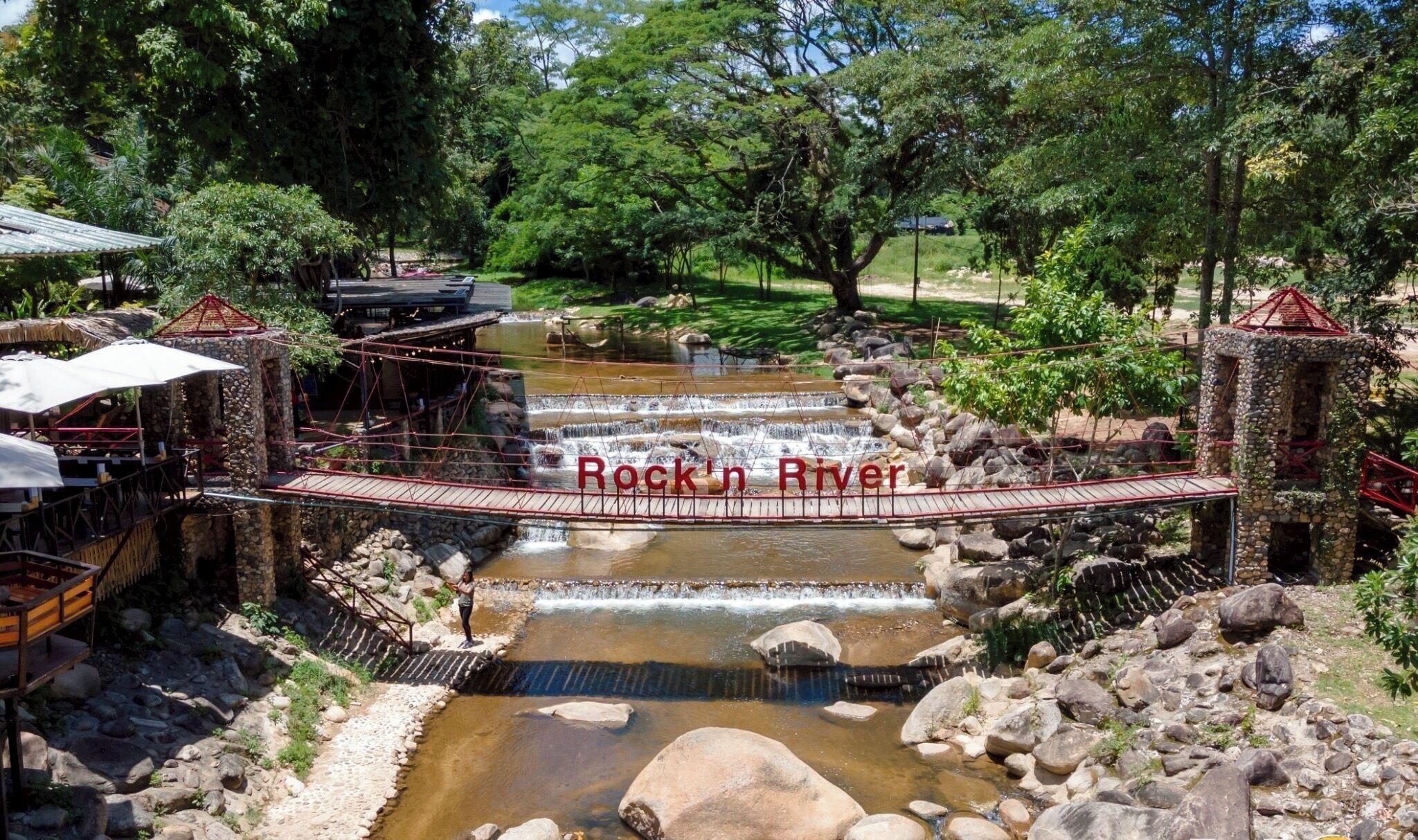 A rustic bridge over a small stream with a wooden deck and metal railings, surrounded by lush greenery and a stone structure.