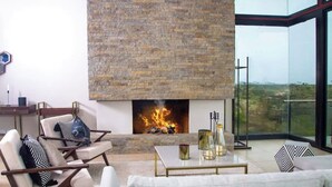 A modern living room with a fireplace, comfortable seating, and a large window offering a view of the outdoors. - Champagne Ridge Villa by YourHost Kenya (Olooloitikosh)