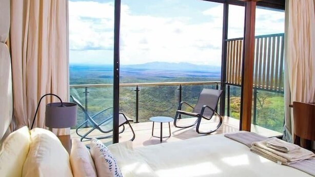 A bedroom with a large bed, a small table, two chairs, and a view of the landscape through large windows. - Champagne Ridge Villa by YourHost Kenya (Olooloitikosh)
