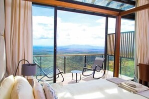 A bedroom with a large bed, a small table, two chairs, and a view of the landscape through large windows. - Champagne Ridge Villa by YourHost Kenya (Olooloitikosh)