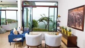 A modern living room with a blue sofa, white armchairs, and a large glass door leading to a balcony with a view of greenery. - Champagne Ridge Villa by YourHost Kenya (Olooloitikosh)