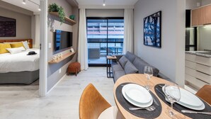 Habitació d'hotel moderna amb un llit, un sofà, zona de menjador amb taula preparada per a dues persones i un gran finestral amb vista a la ciutat.