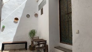 Un estrecho patio con un banco, una mesa y una planta en maceta. Hay platos decorativos en la pared y una ventana con intrincados detalles de metal.