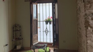Habitación con pared de piedra, techo de madera y vista a través de un arco. Se observa una mesa con un tapete y cojines, una estantería metálica con objetos, y una cesta colgante con flores.