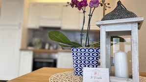 Een keuken met witte kastjes, een blauwe vaas met een patroon en paarse bloemen, een witte lantaarn en een placemat op een houten tafel.