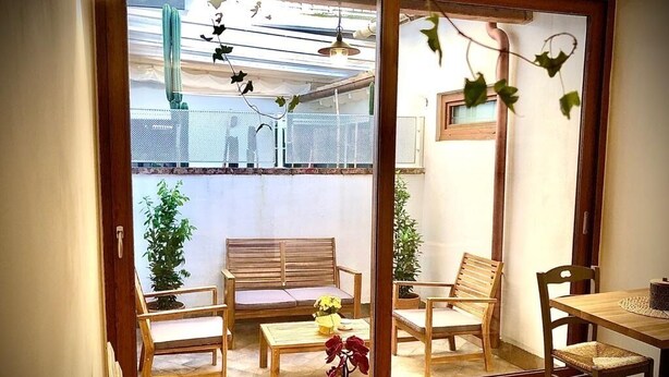 Een kamer met een houten vloer, een bank, twee stoelen, een kleine tafel en een raam met uitzicht op een balkon met planten en een hek.