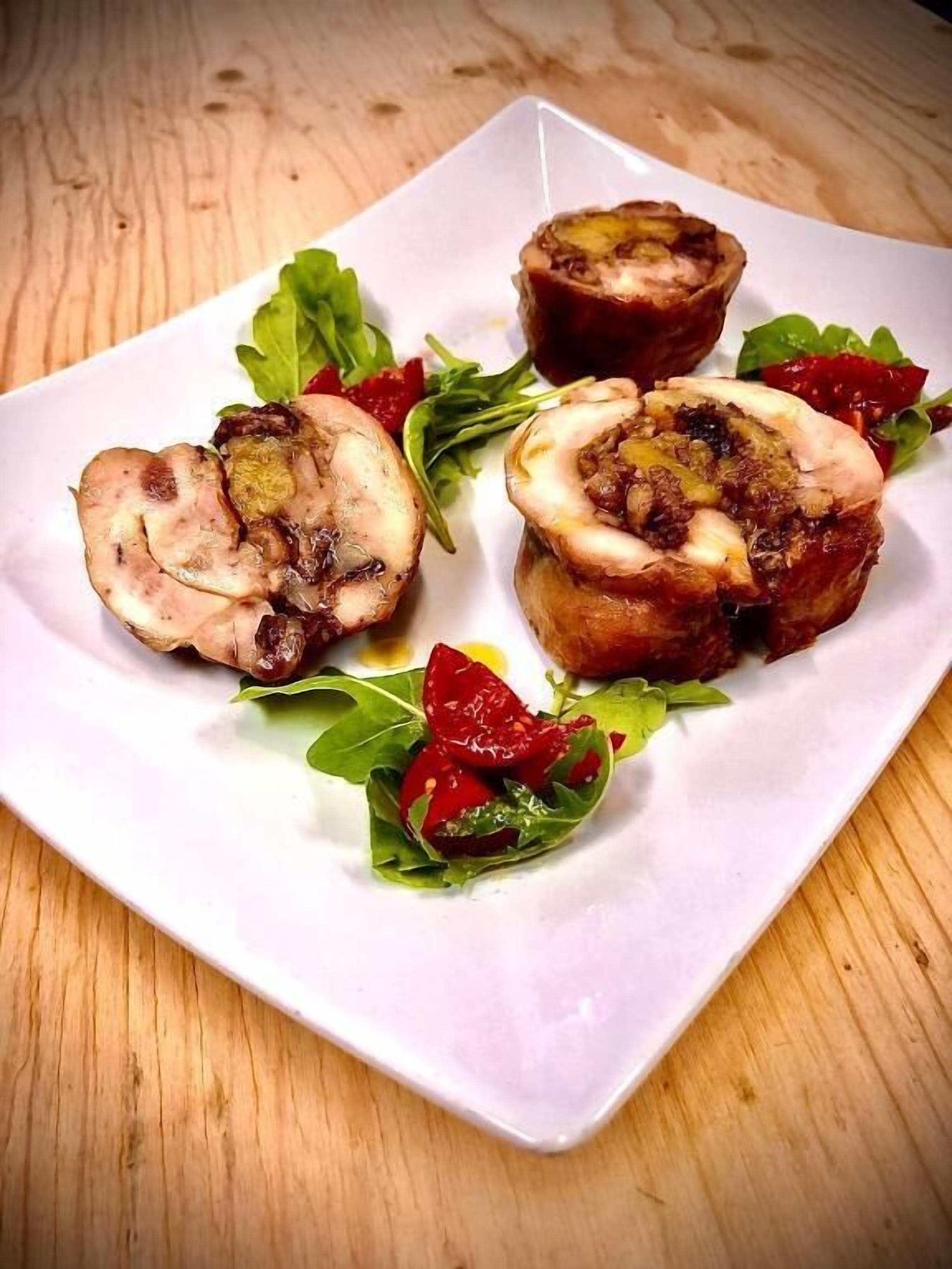 Un plato blanco cuadrado con tres rollos de carne rellenos, adornado con tomates cherry rojos y hojas verdes, sobre una mesa de madera.