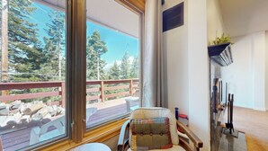 A room with a large window overlooking a deck with a wooden railing and trees. There is a chair with a patchwork cushion and a fireplace with a mantel. - Pet-Friendly Spacious & Bright Home, Mountain Modern. Walk To The Village (Mammoth Lakes)