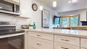 A modern kitchen with a microwave, oven, and a large island with drawers. There is a clock on the wall and a window with a view of trees. - Pet-Friendly Spacious & Bright Home, Mountain Modern. Walk To The Village (Mammoth Lakes)