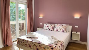 A bedroom with a bed, floral bedspread, two nightstands, a window with curtains, and a view of greenery outside. - ISOLA 1.6.3 Guest House (Fiumicino)