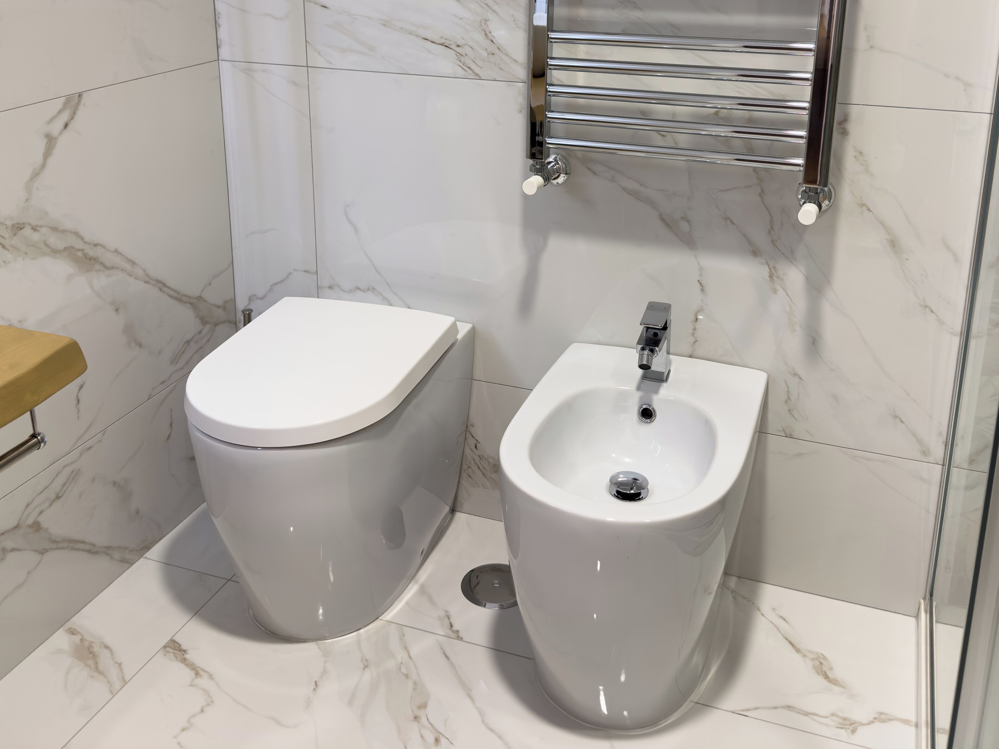 A bathroom with a toilet and bidet, both in white, against a marble-tiled wall.