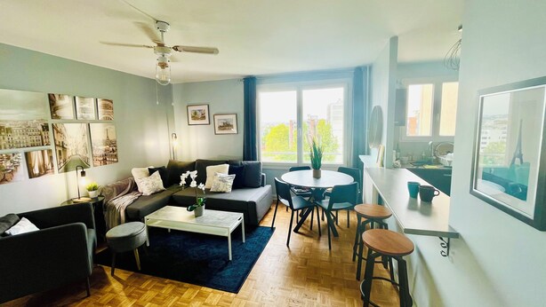 A modern living room with a sofa, coffee table, and a dining area with chairs. There is a ceiling fan, large windows, and artwork on the walls. - Entire rental unit in Maisons-Alfort, France (Maisons-Alfort)