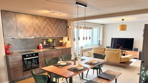 A modern kitchen with a built-in oven, a coffee machine, and a dining area with a wooden table and green chairs. - Elevate Suite 360° (Trier)