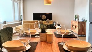 A dining area with a wooden table set for four, surrounded by chairs. There is a television mounted on the wall in the background. - Elevate Suite 360° (Trier)