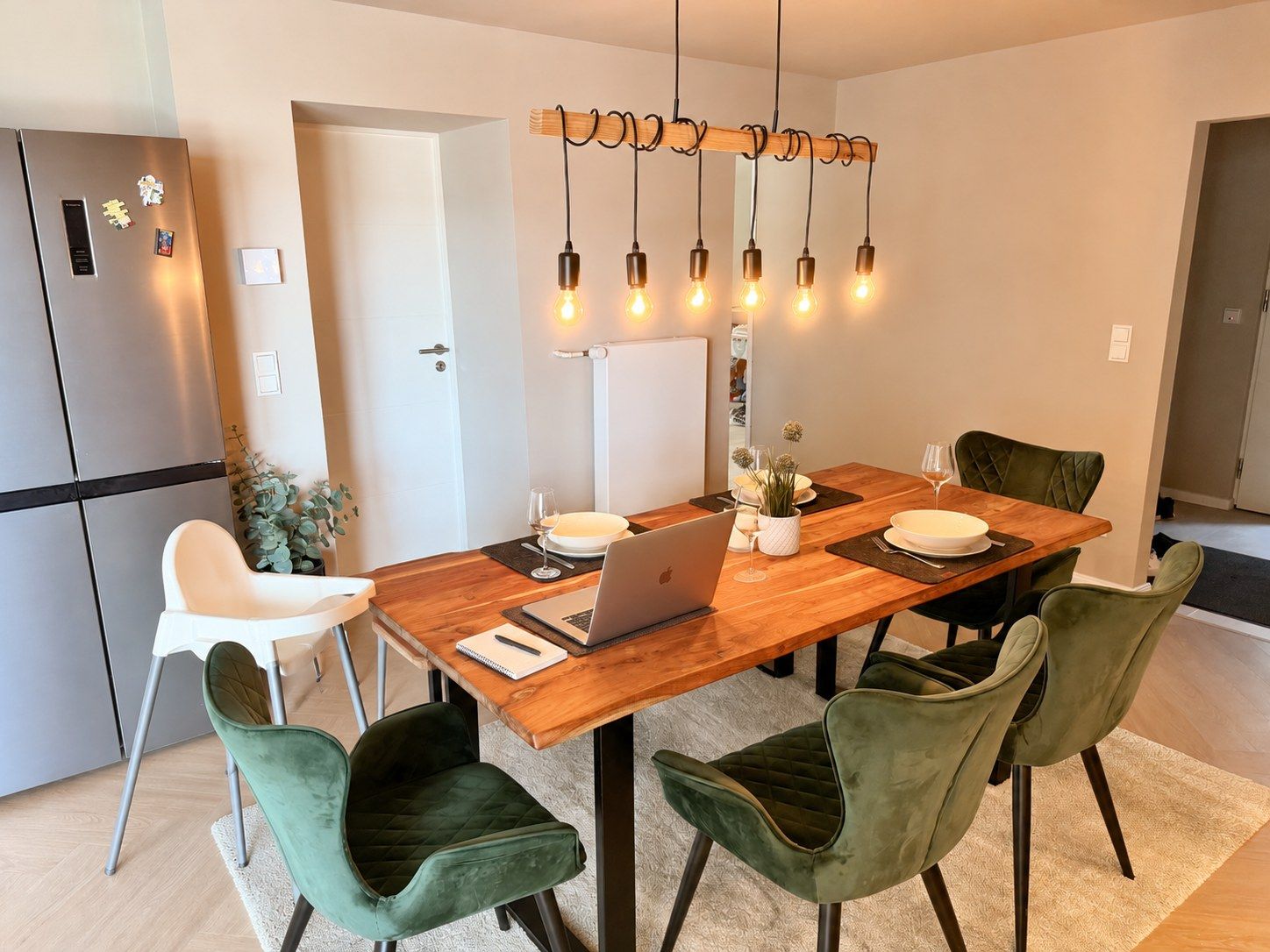 A modern kitchen with a wooden dining table, green upholstered chairs, and a laptop on the table.