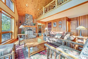 A cozy cabin interior with a stone fireplace, wooden furniture, and a large window overlooking a forest. - Waterfront Lake George Lodge w/ Boat Dock & Deck! (Town of Putnam)