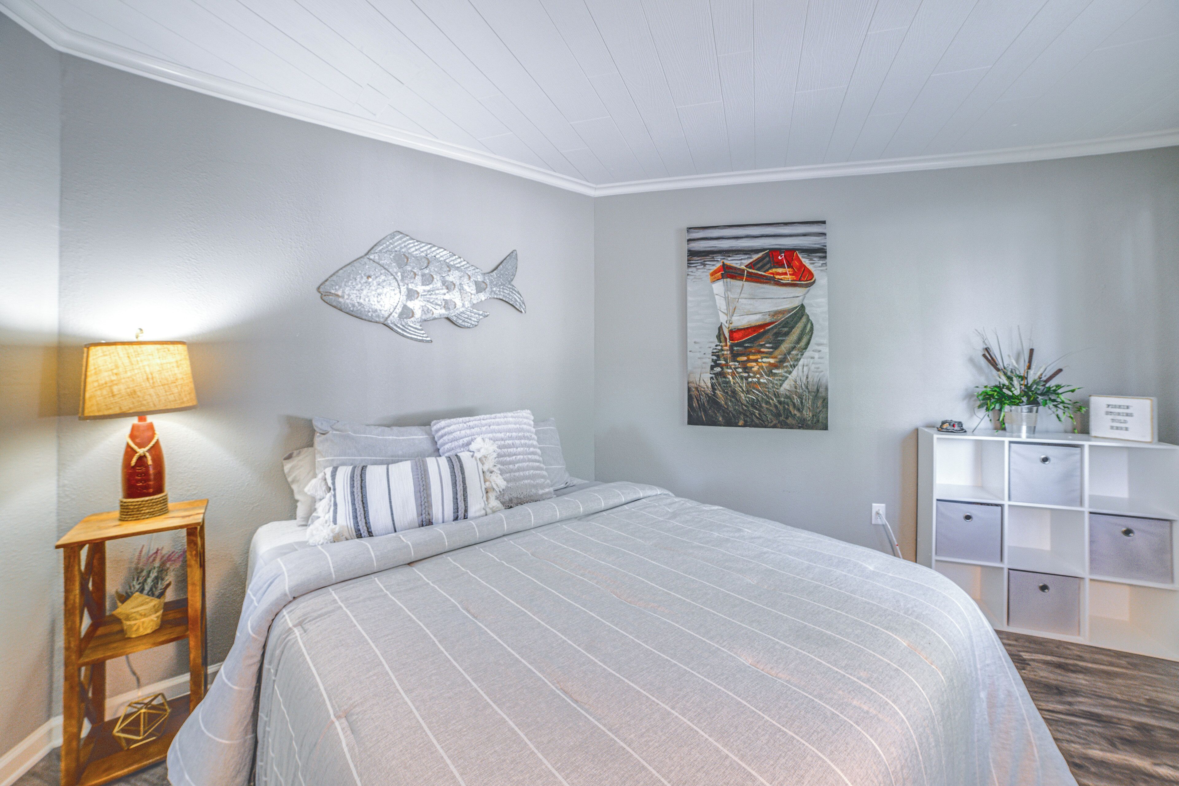 Un dormitorio con cama, un adorno de pez montado en la pared, un cuadro de un barco, una lámpara y un mueble de almacenamiento.