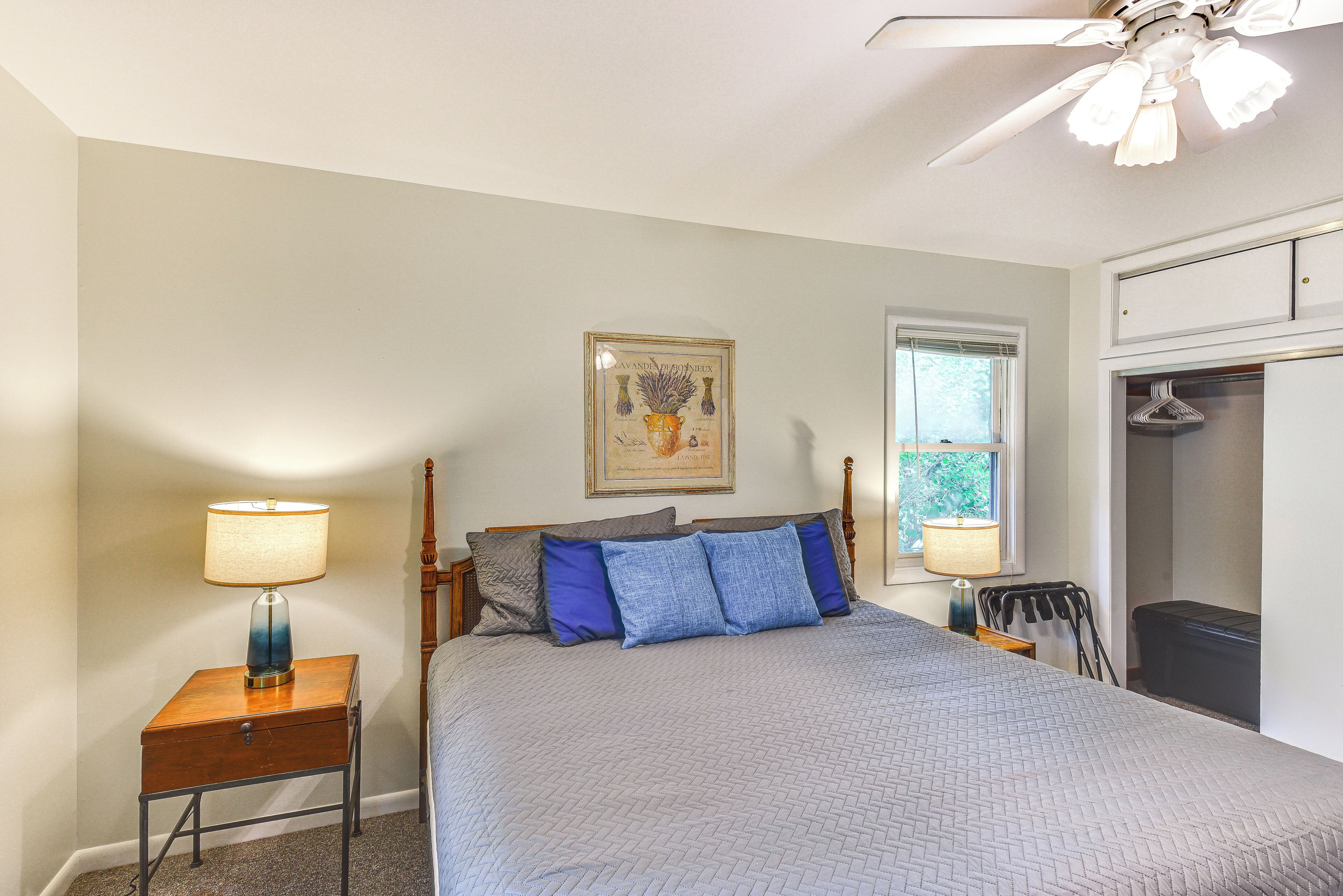 A bedroom with a bed, two bedside tables, a ceiling fan, a window with blinds, and a framed botanical illustration on the wall.