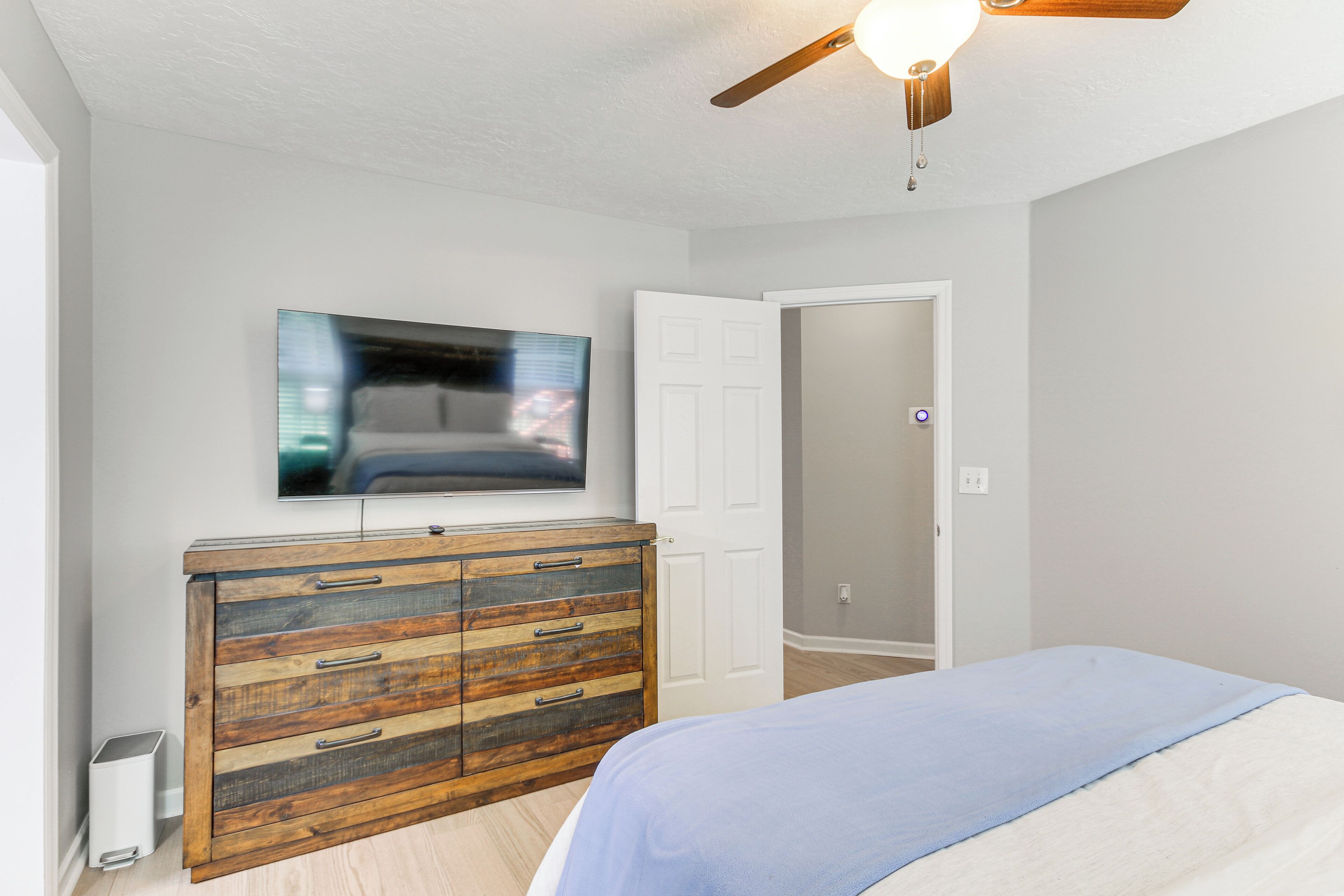 A bedroom with a bed, a wooden dresser, a television on the wall, and a ceiling fan.