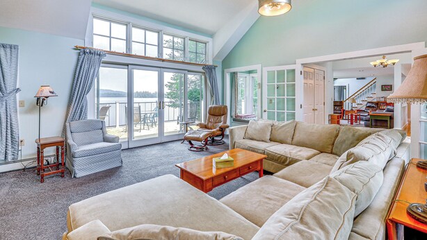 A spacious living room with a large sectional sofa, a rocking chair, and a coffee table. There are large windows providing natural light and a view of the outdoors. - Lakefront New London Retreat w/ Dock & Hot Tub! (New London)