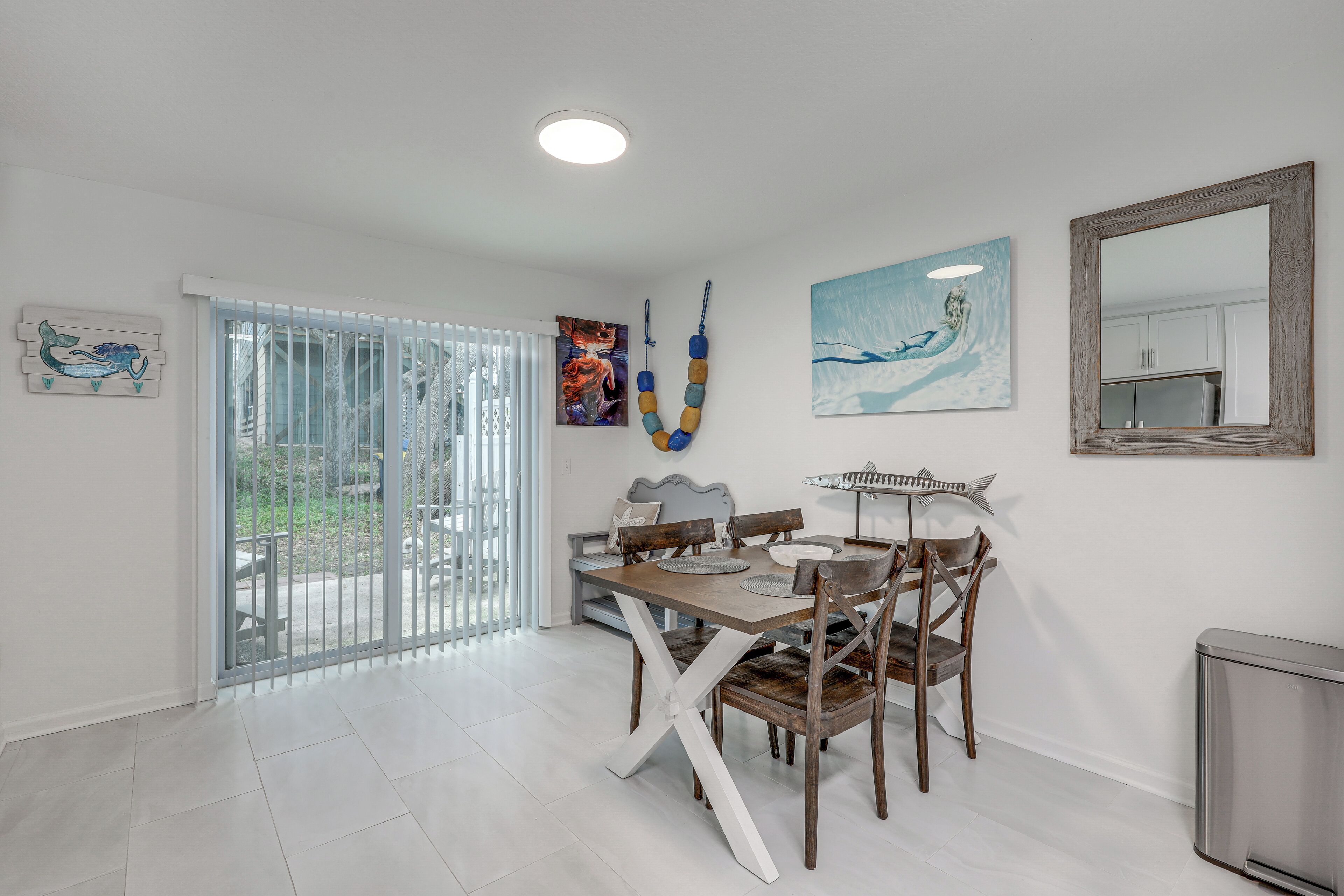 A dining area with a table, chairs, and a bench. There is a window with blinds, a painting, and a mirror on the wall.