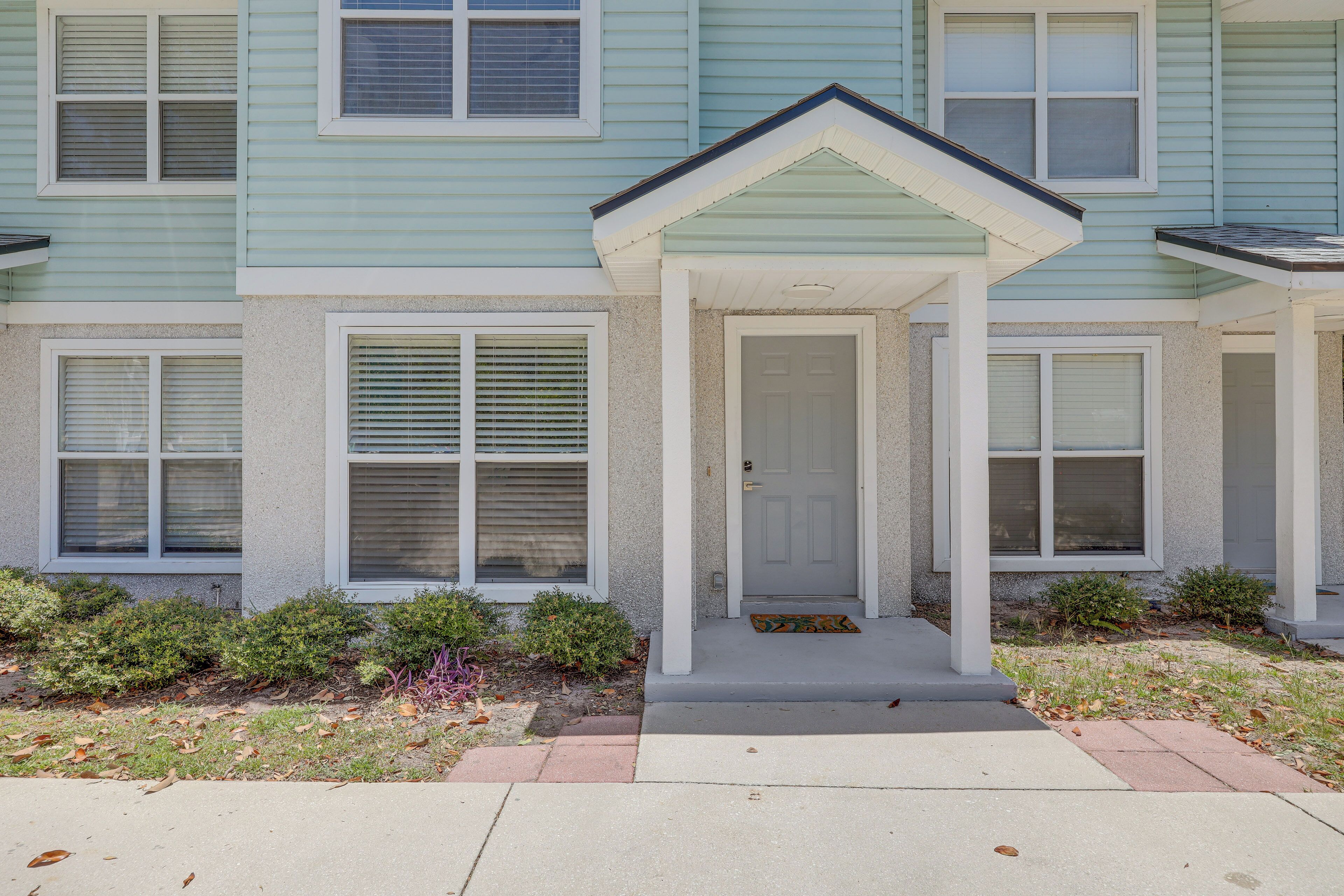 A two-story residential building with a front porch, a door, and a small front yard with grass and shrubs.